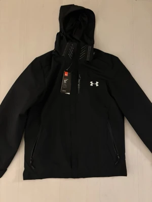 Svart vindjacka från Under Armour - Snygg svart vindjacka från Under Armour med dragkedja framtill, två sidofickor med dragkedja och justerbar huva. Jackan har en diskret logga på bröstet och är tillverkad i ett lätt, vattenavvisande material som passar perfekt för blåsiga dagar. Den går att ta av luva. Kan acceptera vissa prisförslag 