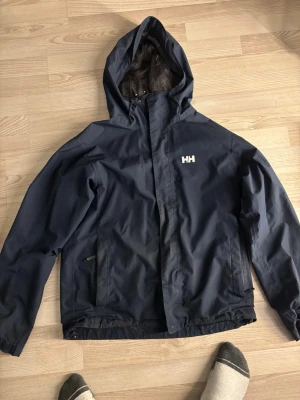 Mörkblå vindjacka från Helly Hansen - Snygg mörkblå vindjacka från Helly Hansen med huva och dragkedja framtill. Jackan har två sidofickor med dragkedja och HH-logga på bröstet. Materialet är slitstarkt och vattenavvisande, perfekt för blåsiga dagar.