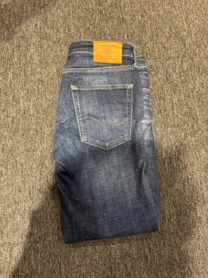 Blå jeans från Jack & Jones - Snygga blå jeans från Jack & Jones med klassisk femficksmodell och ljusa slitningar framtill och baktill. Jeansen har raka ben och är tillverkade i ett mjukt denimtyg som ger en avslappnad look. Perfekta för dig som gillar en tidlös stil. W32 L32