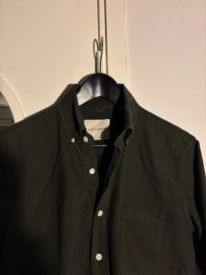 Mörkgrön skjorta från A Day's March - Stilren svart skjorta från A Day's March med klassisk button-down krage och vita knappar. Skjortan har en bröstficka och är långärmad. Tillverkad i mjuk bomull som känns skön mot huden. Perfekt för en clean och avslappnad look.