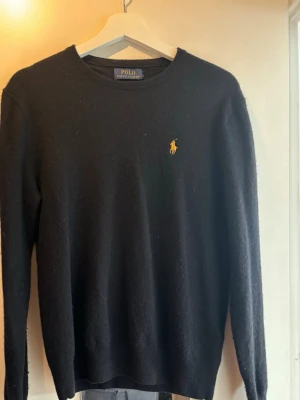 Svart stickad tröja Polo Ralph Lauren - Klassisk svart stickad tröja från Polo Ralph Lauren med rund halsringning och den ikoniska gula broderade loggan på bröstet. Tröjan har långa ärmar och ribbade muddar vid ärmslut och nederkant. Perfekt för en stilren look.