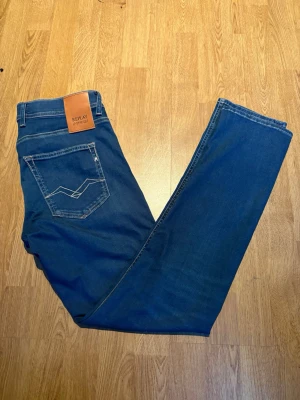 Blå raka jeans från Replay - Snygga blå jeans från Replay med raka ben och klassisk femficksmodell. Jeansen har en cool detalj på bakfickan och läderpatch med logga i midjan. Tillverkade i mjukt denimtyg som sitter skönt hela dagen.