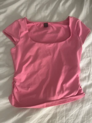 Rosa topp med fyrkantig ringning - Snygg rosa topp med korta ärmar och fyrkantig ringning. Toppens material känns mjukt och stretchigt, perfekt för en trendig look. Passformen är figurnära och den har en enkel, stilren design.