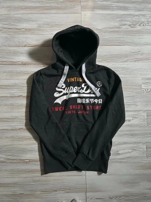 Mörkgrå Superdry hoodie med tryck - Snygg mörkgrå hoodie från Superdry med vit och röd text framtill samt japanska tecken🤩 skön passform! hör gärna av dig vid funderingar samt snabbaffär🙌🏻