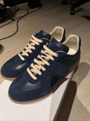 Maison Margiela blå sneakers - Snygga sneakers från Maison Margiela i marinblå mocka och läder med beige snörning och klassisk gummisula. Ikonisk design med låg profil och vita detaljer på insidan. Perfekta för dig som gillar stilrena och trendiga skor.