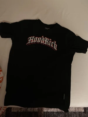 Svart Hoodrich t-shirt med tryck - Säljer en svart t-shirt från Hoodrich med stort vitt och rött tryck på bröstet. Klassisk passform och rund halsringning. Perfekt för dig som gillar streetwear och vill ha en enkel men snygg t-shirt. Priset kan diskuteras eller bytas ut mot nåt