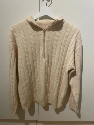 Beige kabelstickad halvzip tröja - Kabelstickad tröja i beige med halvzip och hög krage. Tröjan har långa ärmar och ribbade muddar vid ärmslut och nederkant. Perfekt för lager på lager och ger en klassisk, avslappnad look.