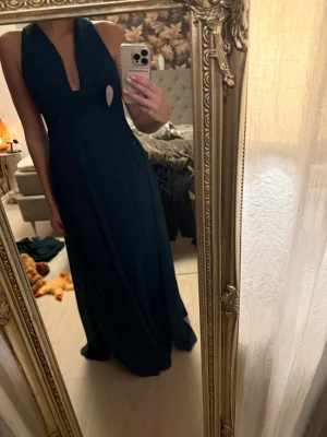 Mörkgrön långklänning med v-ringning - Elegant mörkgrön långklänning med djup v-ringning och korsade axelband i ryggen. Klänningen har en figurnära överdel och ett svepande, lätt utställt kjolparti. Tillverkad i ett mjukt, flytande material som ger en lyxig känsla.