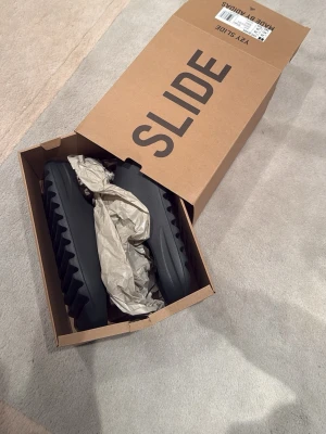 Helt nya Yeezy Slide från Adidas finns kvitto - Säljer ett par svarta Yeezy Slide sandaler från Adidas. De har en chunky, räfflad sula och är tillverkade i ett mjukt, flexibelt syntetmaterial för maximal komfort. Perfekta för en avslappnad och trendig look. Levereras i originalkartong. Små i storlek, helt nya!