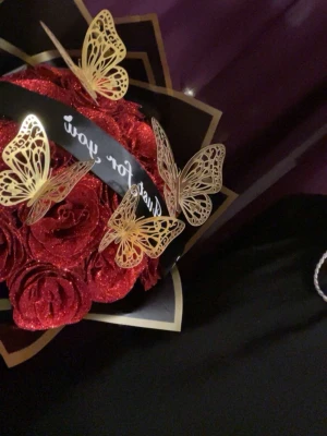 Bukett med röda glittriga rosor - Säljer en dekorativ bukett med konstgjorda röda rosor täckta av glitter, dekorerad med guldfärgade fjärilar i papper och ett svart band med text. Perfekt som present eller dekoration till speciella tillfällen.