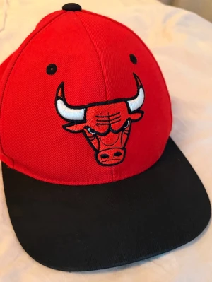 Chicago Bulls keps - Säljer en snygg röd keps med svart skärm från Chicago Bulls. Kepsen har det klassiska Bulls-logotypen broderad framtill och är tillverkad i ett tåligt bomullsmaterial. Perfekt för dig som gillar streetwear och basketstil.
