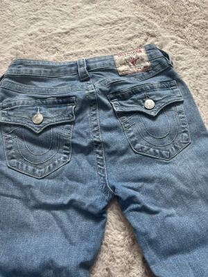 True Religion ljusblå jeans - Snygga ljusblå jeans från True Religion med klassiska fickor bak och silverfärgade knappar. Jeansen har en rak passform och är tillverkade i mjukt denimtyg. Perfekta för en avslappnad och trendig look.