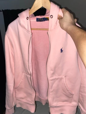 Ljusrosa hoodie från Polo Ralph Lauren - Säljer en ljusrosa hoodie från Polo Ralph Lauren med dragkedja, huva och två stora fickor framtill. Klassisk blå logga broderad på bröstet. Tillverkad i mjukt bomullsmaterial som känns riktigt skönt mot huden. Perfekt för en chill och avslappnad stil. Storlek S säljer denna då jag behöver pengarna. Använd men mycket bra skick