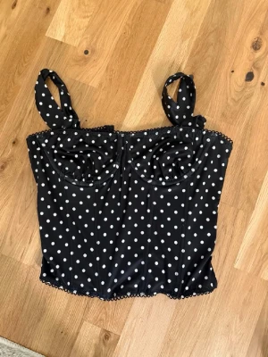 Svart prickig bustier topp - Snygg svart bustier topp med vita prickar och knytbara axelband. Toppen har markerade kupor och en figurnära passform som ger en retro känsla. Perfekt att styla med jeans eller kjol för en trendig look. Materialet känns mjukt och stretchigt.