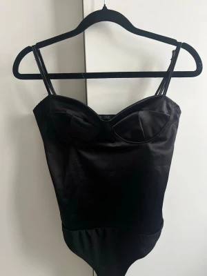 Svart satin body med kupor - Snygg svart body i satin från Nelly med tunna axelband och formade kupor. Materialet har en glansig finish som ger en lyxig känsla och passformen är kroppsnära. 🖤 Passar även storlek XS