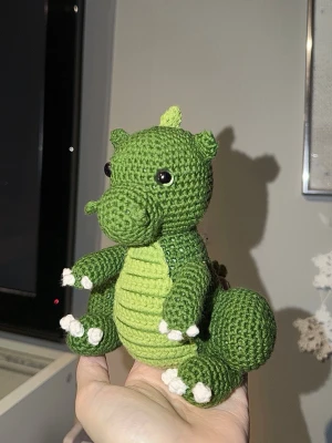 Virkad grön drake  - Handgjord virkad drake i grönt garn med ljusgröna taggar längs ryggen och svansen. Draken har svarta ögon, vit mage och vita klor på tassarna. Tillverkad i bomullsgarn.