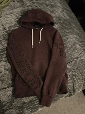 Mörkröd hoodie från Superdry - Säljer en mörklila hoodie från Superdry med broderade detaljer på ärmen och tryck på ena armen. Hoodien har vit dragsko i huvan och mjukt fleecefoder på insidan. Perfekt för dig som gillar streetwear och vill ha något bekvämt och snyggt.