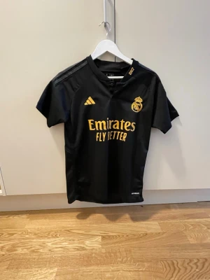 Real Madrid svart matchtröja Adidas - Svart Real Madrid matchtröja från Adidas med guldfärgade detaljer och klubbmärke på bröstet. Tryck med 'Emirates Fly Better' på framsidan. Korta ärmar och sportig passform, perfekt för fotbollsträning eller support.