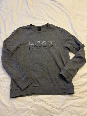 Grå sweatshirt från Hugo Boss - Snygg grå sweatshirt från Hugo Boss med stort BOSS-tryck och broderad logga på bröstet. Tröjan har rund halsringning, långa ärmar och ribbade muddar vid ärmslut och nederkant. Perfekt för en avslappnad och stilren look.
