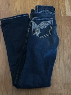 Mörkblå jeans med broderad ficka - Snygga mörkblå jeans från Rock Revival med kontrastsömmar och en broderad bakficka med vinge. Klassisk femficksmodell i jeansmaterial, perfekt för dig som gillar detaljer och streetstil.