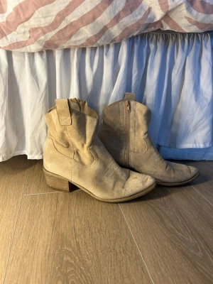 Beige cowboy boots i mocka - Snygga beige cowboy boots i mjuk mocka med klassisk rund tå och låg klack i trälook. Dragkedja på insidan och dekorativa sömmar ger bootsen en autentisk westernkänsla. Perfekta för dig som vill ha en avslappnad men trendig stil.