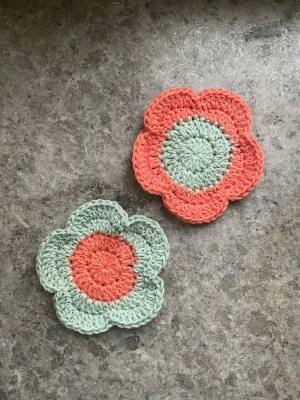 Blommiga virkade glasunderlägg - Två handgjorda glasunderlägg i form av blommor, virkade i bomullsgarn. Ena underlägget är mintgrönt med korallfärgad mitt, det andra är korallfärgat med mintgrön mitt. Perfekta för muggar eller glas och ger en söt retrovibe till köket.