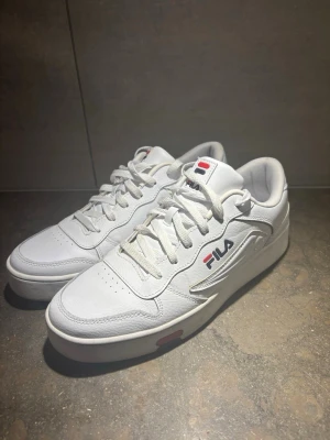 Vita Fila sneakers med logga - Klassiska vita sneakers från Fila med lågt skaft och tydlig Fila-logga i blått och rött på sidan. Skorna har vita snören, diskreta röda detaljer på sulan och är tillverkade i syntetmaterial och läderimitation. Perfekta för en clean och sportig look.