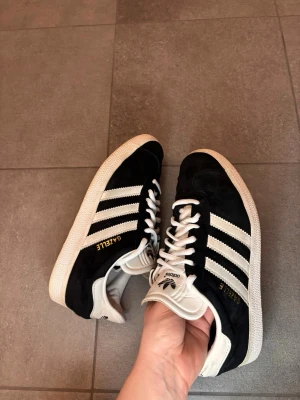 Adidas Gazelle svartvita sneakers - Klassiska Adidas Gazelle sneakers i svart mocka med vita detaljer och de ikoniska tre ränderna på sidan. Skon har vit sula, vita skosnören och guldfärgad Gazelle-text på sidan. Perfekt för dig som gillar retrostil och streetwear.