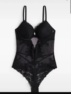 Svart spetsbody med meshdetaljer - Snygg svart body med tunna axelband, vadderade kupor och genomskinliga meshpartier. Dekorerad med vacker spets över hela plagget för en extra lyxig känsla. Perfekt för dig som gillar trendiga och feminina underkläder.