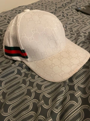 Vit Gucci keps med monogram - Säljer en vit keps från Gucci med det klassiska GG-monogrammet över hela kepsen. På sidan finns en grön och röd rand som ger den en ikonisk look. Kepsen har böjd skärm och justerbar passform. Perfekt för dig som vill ha en stilren och lyxig accessoar. Insidan av kepsen och lite under skärmen har lite smink o sånt.