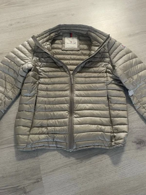 Ljusgrå dunjacka från Moncler - Snygg ljusgrå dunjacka från Moncler med quiltad design och dragkedja framtill. Jackan har ståkrage, två sidofickor och ett unikt serietryck på insidan. Perfekt för kalla vinterdagar och riktigt lätt att bära.