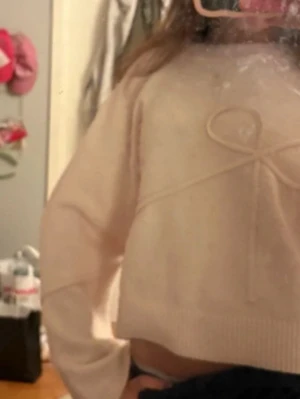 Ljusrosa stickad tröja med rosett - Supersöt ljusrosa stickad tröja med en stor rosettdetalj framtill. Tröjan har lång ärm, croppad passform och ribbade muddar vid ärmslut och nederkant. Perfekt för dig som gillar gulliga detaljer och en mjuk känsla.