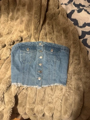 Blå croppad jeansbustier - Säljer en blå croppad bustier i jeans med knappar framtill och rå, fransig kant nedtill. Toppen har dekorativa bröstfickor och smockad rygg för skön passform. Perfekt för dig som vill ha en edgy och trendig look.
