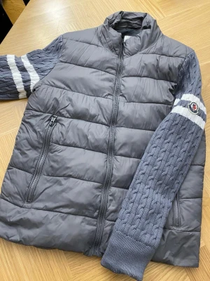 Moncler mörkgrå pufferjacka med stickade ärmar - Moncler pufferjacka i mörkgrått med stickade, kabelmönstrade ärmar och vita ränder. Jackan har dragkedja fram och på fickorna, samt Moncler-logga på ärmen. Kroppen är quiltad och fodrad, perfekt för kyliga dagar.