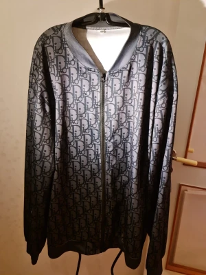 Svart bomberjacka med monogram - Snygg svart bomberjacka med heltryckt monogrammönster i grått. Jackan har dragkedja framtill, ribbade muddar och krage i blankt material. Perfekt för dig som gillar streetwear och vill sticka ut med en unik design.