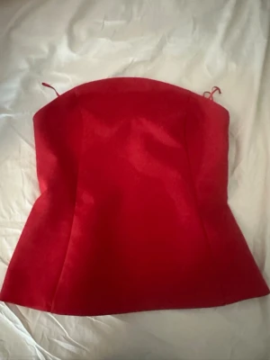 Röd axelbandslös korsettopp - Snygg röd korsettopp med axelbandslös design och lätt utsvängd nederkant. Perfekt för att skapa en statement-look och matcha med både jeans och kjol. Toppens material har en slät finish och ger en elegant siluett.