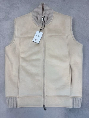 Beige dunväst från Gran Sasso - Stilren beige dunväst från Gran Sasso med mjukt teddyfoder och ribbstickad hög krage. Västen har dragkedja framtill och diskreta sidofickor. Perfekt för lager på lager och ger en clean look till din outfit.