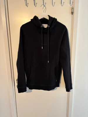 Les Deux hoodie   - Snygg svart hoodie från Les Deux med klassisk känguruficka och justerbar huva med snören. Diskret broderad logga på bröstet. Tillverkad i mjukt bomullsmaterial som känns skönt mot huden. Perfekt för en avslappnad och stilren look.