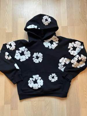 Svart hoodie med vita blommor - Snygg svart hoodie med stora vita blommönster över hela tröjan. Hoodien har en klassisk huva och en stor magficka framtill. Perfekt för dig som vill sticka ut med en unik och trendig stil. Mjuk och skön, passar till alla tillfällen.
