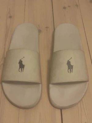 Vita tofflor från Polo Ralph Lauren - Säljer ett par vita slides från Polo Ralph Lauren med den klassiska polospelaren tryckt på ovandelen. Tofflorna har en enkel och stilren design med bred rem och platt sula, perfekta för chill dagar hemma under sommaren eller på stranden.