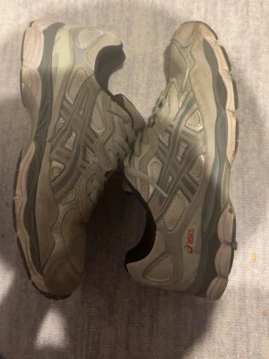 gråa Asics sneakers med meshdetaljer - Säljer ett par gråa sneakers från Asics med grå och röda detaljer. Skorna har meshpaneler för extra ventilation och klassiska Asics-loggor på sidorna. Tjock sula för bra komfort och snörning framtill. Perfekta för dig som gillar sportig stil.