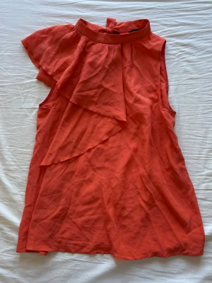 Röd ärmlös blus med volang - Snygg orange/korall blus med volangdetalj framtill och knytning i nacken. Ärmlös modell med asymmetrisk volang 