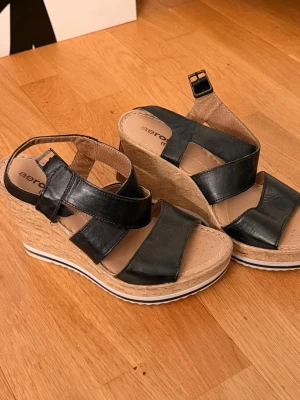 Svarta kilklackssandaler från Aeros me - Säljer ett par svarta sandaler från Aerosoft med breda remmar i konstläder och super bekväm kilklack i korkimitation. Storlek 37