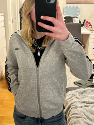Grå Adidas hoodie med dragkedja - Säljer en grå hoodie från Adidas med dragkedja framtill och svarta klassiska ränder längs ärmarna. Tröjan har huva och fickor på sidorna. Perfekt för en sportig och avslappnad stil. Materialet är mjukt och bekvämt.