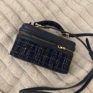 Svart tweed handväska med gulddetaljer - Snygg svart handväska i tweed och konstläder med guldfärgade dragkedjor och detaljer. Väskan har både ett handtag och en lång axelrem, samt en praktisk ficka på baksidan. Perfekt för dig som gillar en trendig och stilren look.