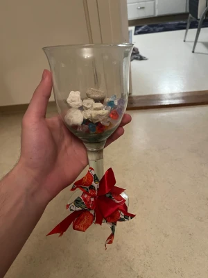 Dekorativt glas med stenar och pärlor - Stort vinglas fyllt med vita och beige stenar samt färgglada plastpärlor i blått, rött och lila. Glaset är dekorerat med ett rött band och mönstrad rosett runt foten. Perfekt som dekoration till rummet eller present.