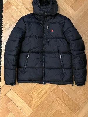 Svart pufferjacka från Polo Ralph Lauren - Svart pufferjacka från Polo Ralph Lauren med huva och tvåvägsdragkedja. Perfekt för kalla dagar. Köpt här på Plick, men passade inte.|                                                                     