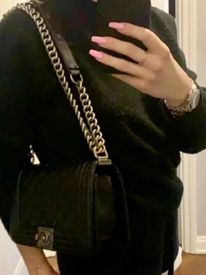 Svart fake Chanel Boy Bag i läder/ Quilted Medium Le Boy - Säljer en fake svart Chanel Boy Bag i quiltat läder med silverfärgad kedjerem och ikoniskt CC-spänne framtill. Väskan har en boxig form och coola detaljer som ger en lyxig vibe. .