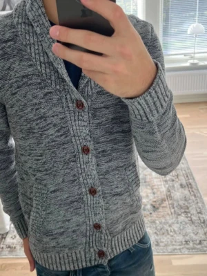 Grå cardigan Pier One - Snygg gråmelerad kofta med sjalkrage och bruna knappar. Koftan har ribbade muddar vid ärmslut och nederkant samt två fickor framtill. Perfekt för lager på lager och chill stil. Materialet är mjukt och stickat för extra komfort.