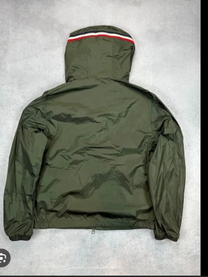 Grön vindjacka från Moncler - Stilren mörkgrön vindjacka från Moncler med huva och dragkedja framtill. Jackan har elastiska muddar, Moncler-logga på ärmen och snygga rödvitblå detaljer på huvan. Perfekt för blåsiga dagar och enkel att matcha med olika outfits.